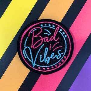 May include: A black and pink embroidered patch with the text "Bad Vibes" in a retro neon sign style.