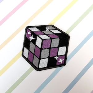 Peut inclure: Un patch brodé représentant un cube 3D avec un motif d'étoile, en noir, blanc, gris et violet.