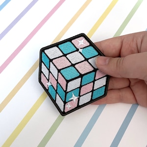 May include: A black and white embroidered patch of a 3D cube with pink, blue, and white squares. The cube has a glittery effect.