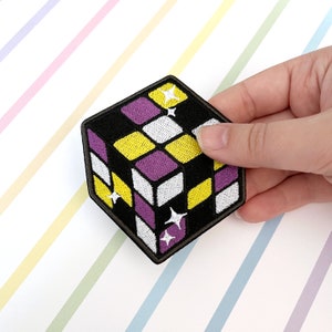 May include: A black, white, yellow and purple embroidered patch shaped like a cube with a glittery design. The patch is a representation of the non-binary gender identity.