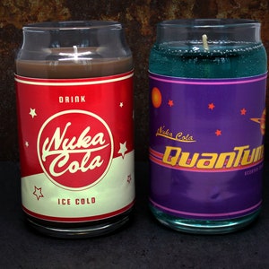 May include: Two glass candle jars shaped like soda cans. The left jar has a red and white label with the text "Drink Nuka Cola Ice Cold". The right jar has a purple label with the text "Nuka Cola Quantum".