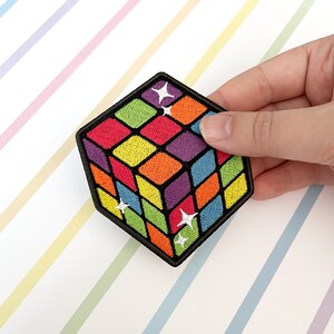 May include: A colourful embroidered patch shaped like a cube with a rainbow pattern and white stars. The patch is perfect for adding a pop of colour to clothing, bags, or other accessories.