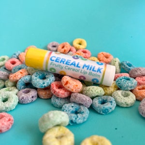 May include: A white lip balm tube with the text "Cereal Milk Fruity Cereal Lip Balm" on a blue background surrounded by colorful cereal rings.