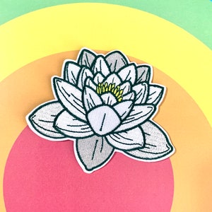 Puede incluir: Un parche de hierro blanco bordado de una flor de nenúfar con contornos verdes y un centro amarillo.