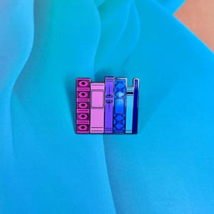 May include: A colourful enamel pin depicting a stack of books with different coloured spines. The books are pink, purple, blue, and light blue. The pin is a great way to show your love of reading.