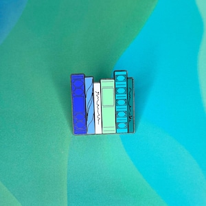 May include: A colourful enamel pin featuring a stack of five books with different coloured spines. The books are blue, white, green, and turquoise.