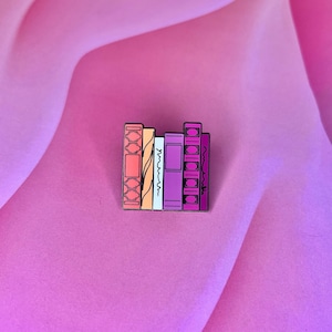 May include: A colorful enamel pin featuring a stack of five books with different colored spines. The books are in shades of pink, orange, yellow, blue, and purple.