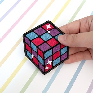 May include: An embroidered iron-on patch depicting a 3D cube with a geometric pattern of red, blue, and purple squares. The cube has white stars on each side.