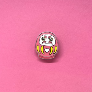 May include: A pink and yellow enamel pin of a traditional Japanese daruma doll with a white face and black eyes. The doll has a yellow heart on its chest and yellow stripes around its body.
