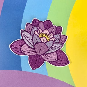 Könnte beinhalten: Lilafarbener gestickter Blumen-Patch mit pinkfarbenem Zentrum und gelben Akzenten. Die Blume hat ein detailliertes Design und ist perfekt, um jedem Projekt einen Hauch von Natur zu verleihen.