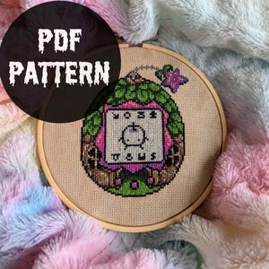 Könnte beinhalten: Ein Kreuzstichmuster in einem Holzrahmen mit dem Text "PDF PATTERN". Das pixelige Design zeigt einen Retro-Videospielcharakter in Grün, Rosa und Braun. Der Hintergrund ist ein weicher, mehrfarbiger Stoff.