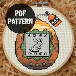 Könnte beinhalten: Ein Kreuzstichmuster in einem Holzrahmen mit einem pixeligen Design eines Kaninchens, einer Flasche mit brennender Lunte und dem Text "PDF PATTERN". Das Design verwendet Braun-, Grün-, Orange- und Weißtöne.