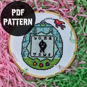 Puede incluir: Patrón de punto de cruz con un diseño de juego portátil retro, en un aro de bordado de madera. El diseño presenta un personaje pixelado. El texto "PDF PATTERN" está en un círculo negro. Fondo de papel triturado rosa y verde.