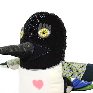 May include: A handmade plush bird with a black head and beak, white body, and patterned wings. The bird has large, expressive eyes with yellow and black details. A pink heart is sewn on the front.