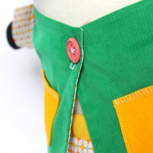 May include: Close-up of a colorful fabric item with a green top, yellow pockets, and a pink button. The item has a white lining and a patterned fabric peeking out from underneath. The stitching is white.