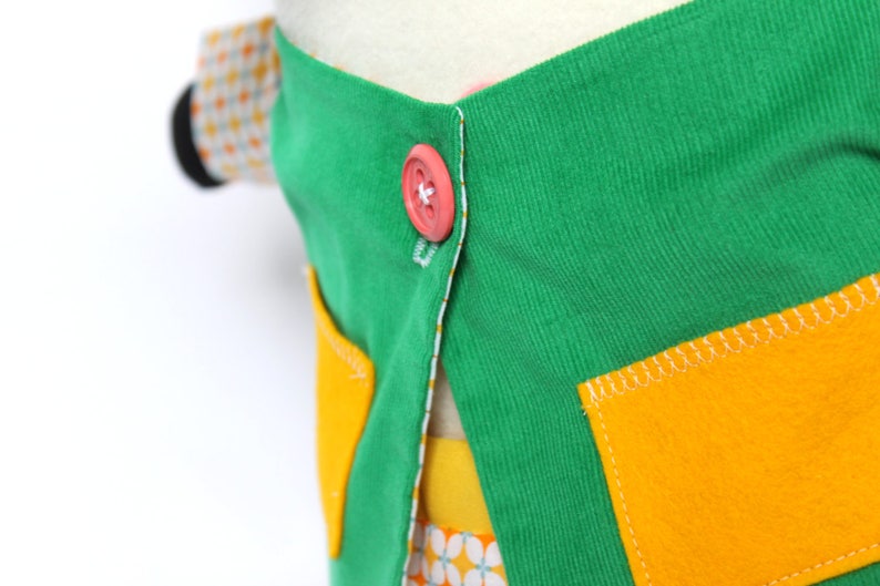 May include: Close-up of a colorful fabric item with a green top, yellow pockets, and a pink button. The item has a white lining and a patterned fabric peeking out from underneath. The stitching is white.