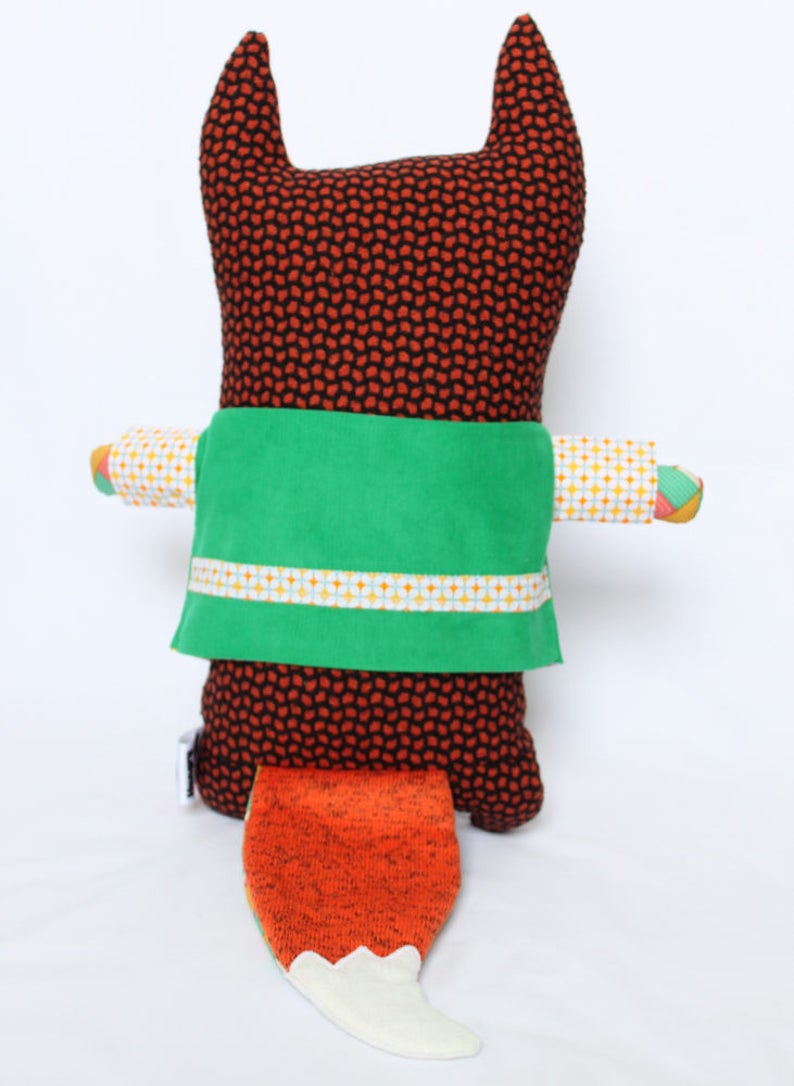 May include: A handmade stuffed fox doll with a brown body covered in small orange and black dots. It has a green sash with a white and yellow patterned trim. The fox's tail is orange with a white tip. The arms are patterned with yellow, white, and orange.