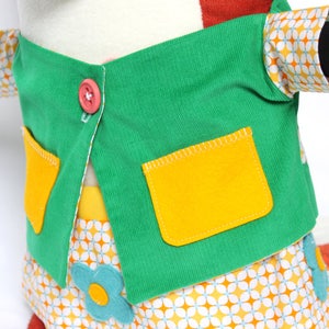 May include: A plush toy wearing a green jacket with yellow pockets and a pink button. The sleeves have a retro floral pattern. The skirt has a matching floral pattern with blue flower accents. The toy's arms have black cuffs.