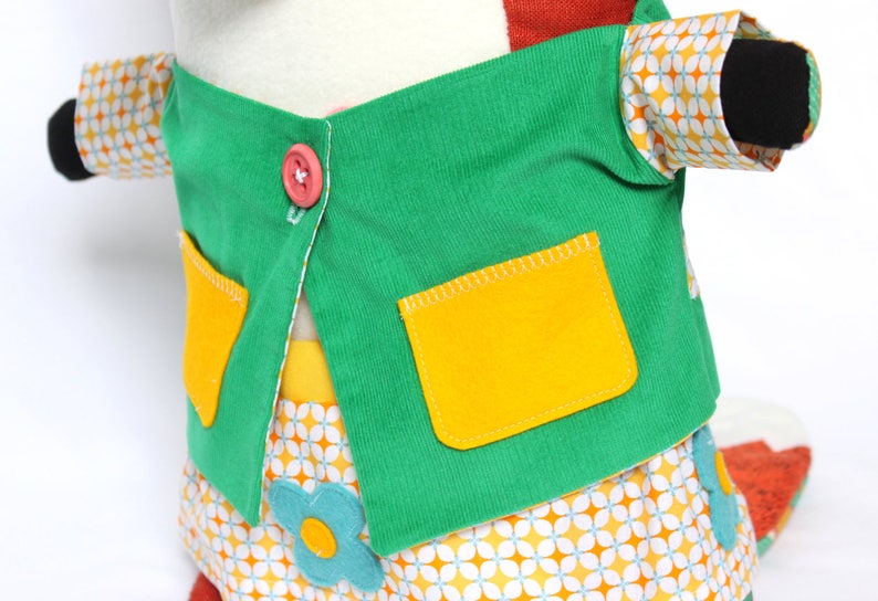 May include: A plush toy wearing a green jacket with yellow pockets and a pink button. The sleeves have a retro floral pattern. The skirt has a matching floral pattern with blue flower accents. The toy's arms have black cuffs.