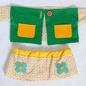 May include: A doll outfit set featuring a green vest with yellow pockets and a pink button, and a yellow skirt with a floral pattern and turquoise flower accents. The outfit has a retro aesthetic.