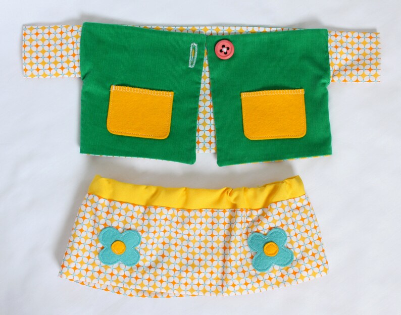 May include: A doll outfit set featuring a green vest with yellow pockets and a pink button, and a yellow skirt with a floral pattern and turquoise flower accents. The outfit has a retro aesthetic.