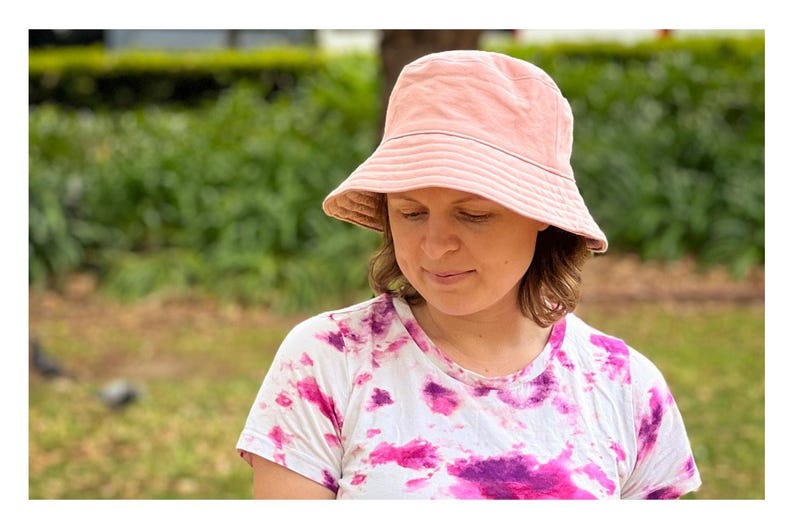 May include: A woman wearing a pink bucket hat with a white tie-dye shirt. The hat has a white band around the crown.