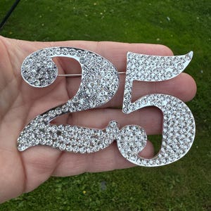 May include: A silver-toned brooch in the shape of the number 25, completely covered in clear rhinestones.