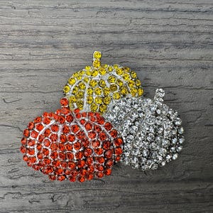 May include: Three jeweled pumpkin brooches in red, yellow, and white. The brooches are made of rhinestones and are perfect for fall fashion.