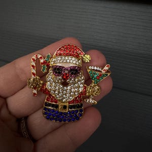 May include: A Christmas-themed brooch depicting Santa Claus. The brooch has a gold-tone finish and is adorned with red, white, and blue rhinestones. Santa wears purple sunglasses and holds a candy cane and a cocktail. The brooch is a festive accessory.