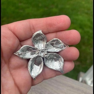 Pu&ograve; includere: Broche a forma di fiore argentato con chiusura a spilla. Il fiore ha cinque petali e un centro con un design in rilievo.