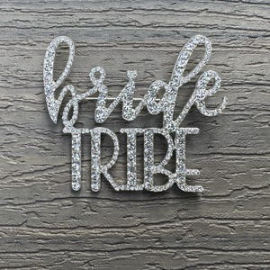 May include: Silver rhinestone bridal party pin with the words "bride tribe" in a cursive font.
