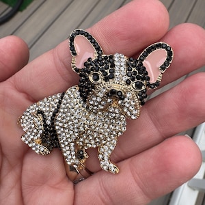 Puede incluir: Broche de bulldog francés en tono dorado, incrustada con cristales transparentes y negros. El perro tiene orejas negras, cara negra y blanca y cuerpo dorado. Accesorio decorativo.