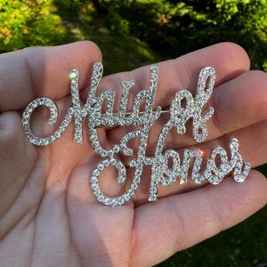 May include: A silver-toned brooch with the words "Maid of Honor" spelled out in rhinestones.