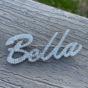 May include: A silver-toned brooch with the word "Bella" spelled out in rhinestones.