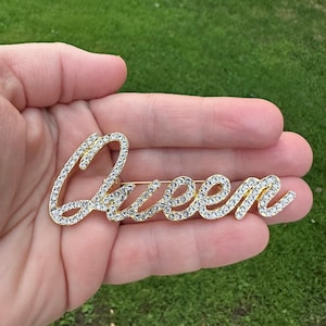 Peut inclure: Broche dorée avec des strass clairs épellant le mot "Queen".