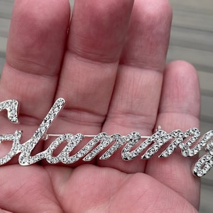 May include: A silver-toned brooch spelling "Glammy" in cursive script, encrusted with sparkling rhinestones. The brooch has a pin backing for secure attachment. A stylish accessory for adding a touch of glamour to any outfit.