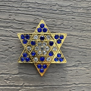 Puede incluir: Un broche dorado con forma de Estrella de David, adornado con piedras preciosas azules y transparentes. El broche presenta un diseño simétrico de estrella de seis puntas, con las piedras preciosas engastadas en las puntas y el centro de la estrella. El broche se muestra sobre una superficie gris texturizada.