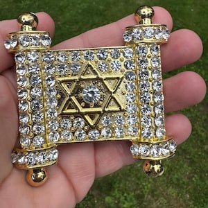 May include: A gold-colored, scroll-shaped pendant encrusted with clear rhinestones. The pendant features a Star of David design in the center and is held in a hand against a green background. The scroll ends have a rounded shape.