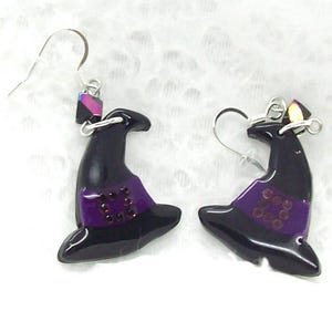 May include: A pair of dangle earrings featuring black witch hat charms with purple bands and small dark red circular accents. The earrings are attached to silver-toned metal hooks and include a small iridescent geometric bead above each hat.