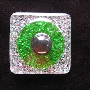 Può includere: Un fermacarte quadrato in vetro trasparente con un centro di glitter verde e un cerchio di metallo argentato al centro.