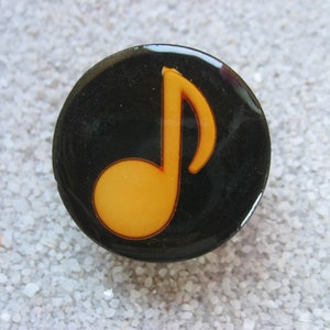 Può includere: Uno spillo a forma di nota musicale nero e arancione. La nota è di un arancione brillante e su uno sfondo nero.