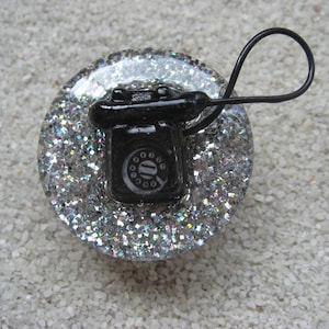 Könnte beinhalten: Ein schwarzer, telefonförmiger dekorativer Knopf mit Wählscheibe sitzt auf einer silbernen, glitzerbedeckten Basis. Der Knopf hat ein schwarzes Kabel. Der Hintergrund ist eine hellfarbige, strukturierte Oberfläche.