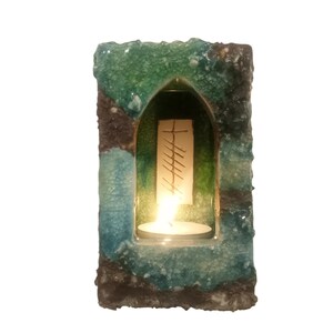 May include: A teal blue and green ceramic candle holder with a small window and a lit tea light inside. The window has a white background with a brown inscription.