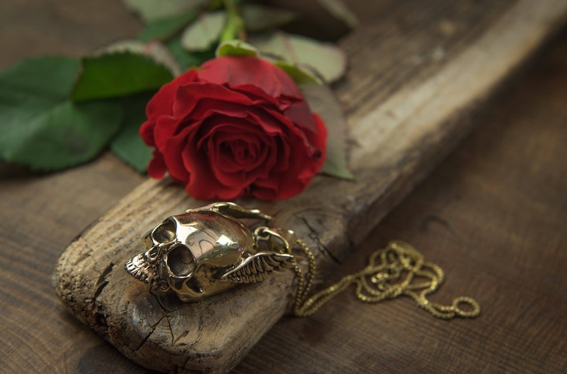 May include: A gold skull pendant with wings on a gold chain, resting on a piece of weathered wood with a red rose.