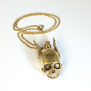 May include: A gold-tone necklace with a skull pendant. The skull has wings and is detailed with a textured surface.