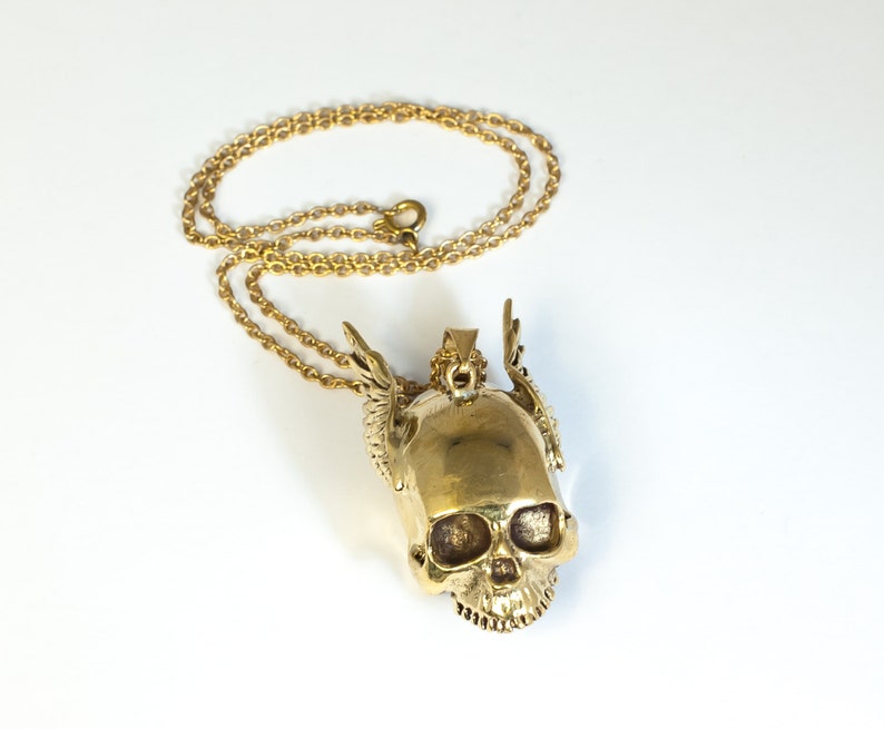 May include: A gold-tone necklace with a skull pendant. The skull has wings and is detailed with a textured surface.
