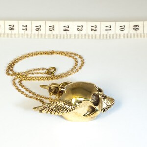 May include: A gold-colored necklace with a winged skull pendant. The pendant is detailed with a skull face and wings.