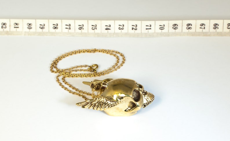 May include: A gold-colored necklace with a winged skull pendant. The pendant is detailed with a skull face and wings.