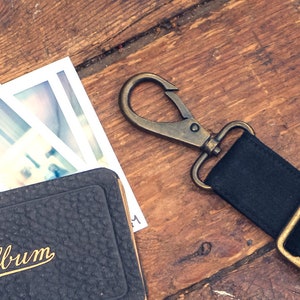 May include: A black leather photo album with the word "Album" written in gold script on the front. The album is closed and is sitting on a wooden surface. There are three instant photos on top of the album. A black canvas strap with a brass clasp is also on the surface.