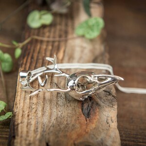 May include: Silver pendant necklace featuring a detailed bird skull with antlers. The necklace is on a wooden surface with green leaves.
